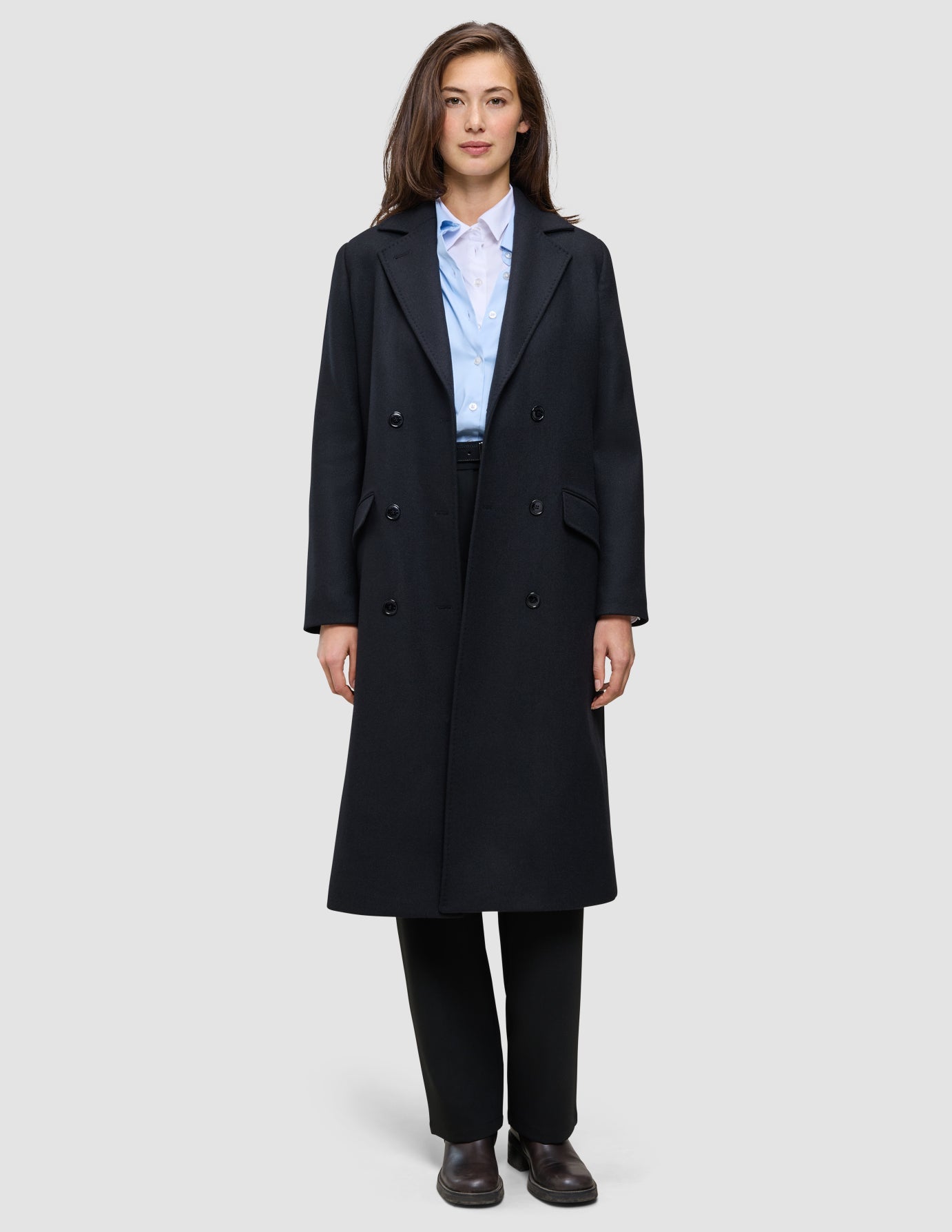 Eine Frau trägt einen schwarzen Damen-Wollmantel in knielanger Länge im Zweireiher-Stil über einem hellblauen Hemd mit Knöpfen und einer schwarzen Hose, was das klare skandinavische Design unterstreicht.